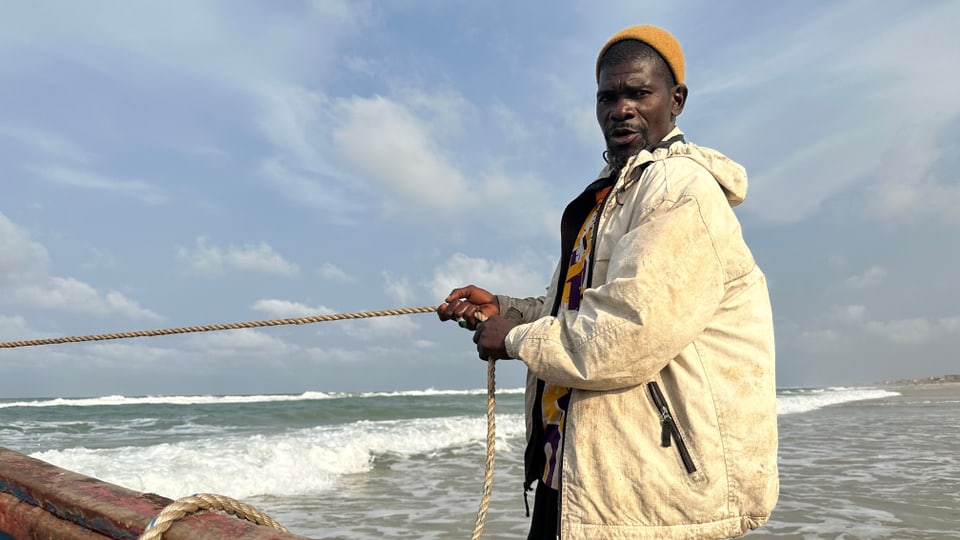 Moussa Cissé hantiert mit einem Seil an einem Strand und blickt in die Kamera