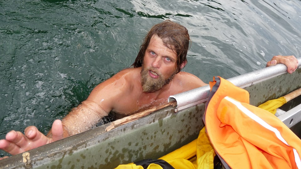 Mann mit langen Haaren und Bart im Wasser, hält sich an Boot fest