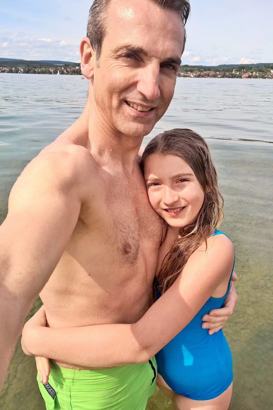 Vater und Tochter am Ufer eines Sees in Badehosen. Es scheint kalt zu sein.