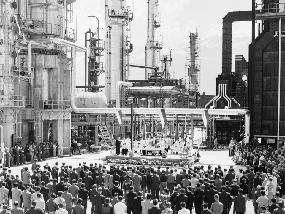 Schwarz-Weiss-Bild, zahlreiche Menschen stehen vor den Türmen der Raffinerie. Auf einer kleinen Bühne steht ein Priester