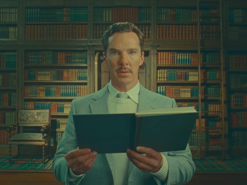 Ein Mann mit Schnurrbart steht in einer Bibliothek. Er hält ein Buch in den Händen