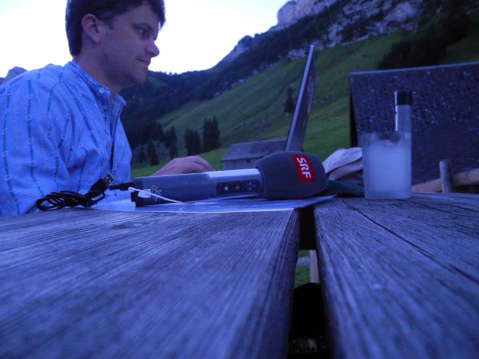 Reporter Roman Portmann mit Microfon und Laptop auf hölzernem Tisch im Freien vor der Alphütte der Fässlers.