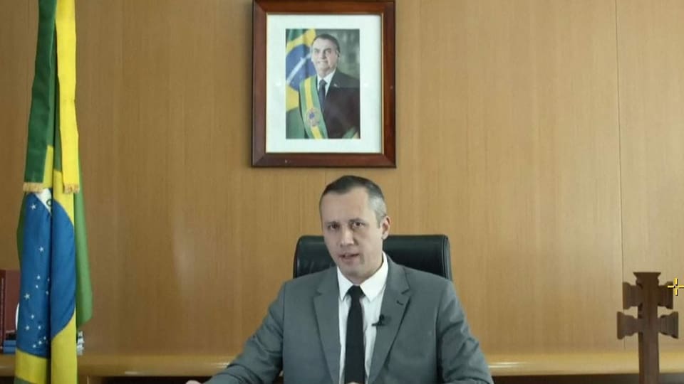 Mann sitzt an Pult, dahinter Bild von Bolsonaro. Auf dem Pult steht ein Holzkreuz.