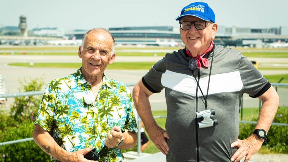 Zwei ältere Männer, einer kleiner, der andere mit blauer Mütze, vor dem Flugplatz.