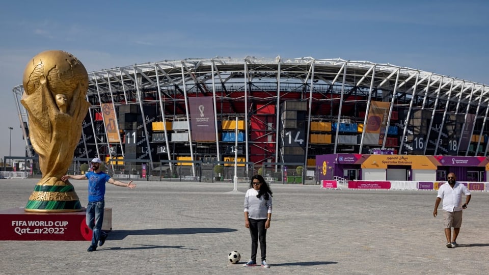 Zwei Männer und eine Frau (ohne Kopftuch) stehen vor einem Stadion, welches mit bunten Plakaten geschmückt ist.