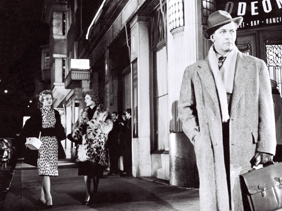 Ein Mann mit Mantel, Hut und Aktentasche steht auf der Strasse vor dem Café Odeon. Zwei elegant gekleidete Damen schlendern auf dem Bürgersteig entlang.