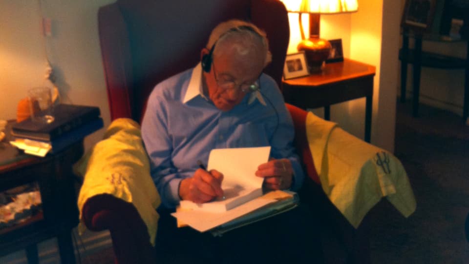Werner Kleeman sitzt in einem Sessel und schreibt eine Widmung in ein Buch.