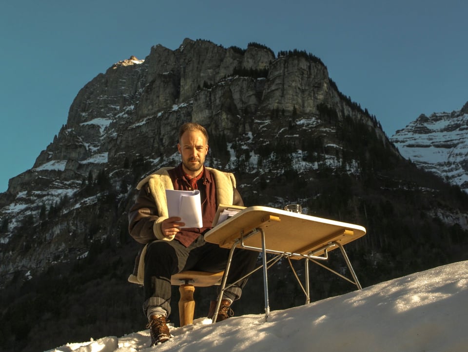 junger Mann auf Holzschemel im Schnee, dahinter ein Berggipfel, vor ihm ein Tisch, in der Hand ein Heft.