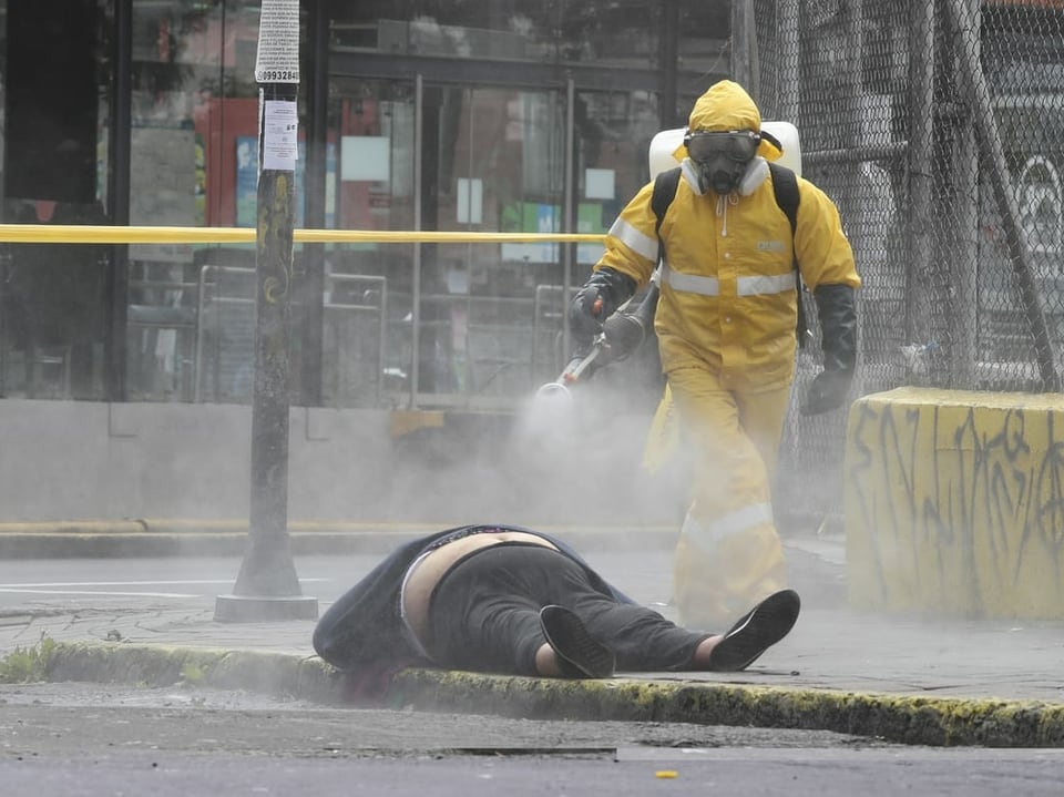 Person in Schutzanzug besprüht eine am Boden liegende Leiche mit Desinfektionsmittel. 