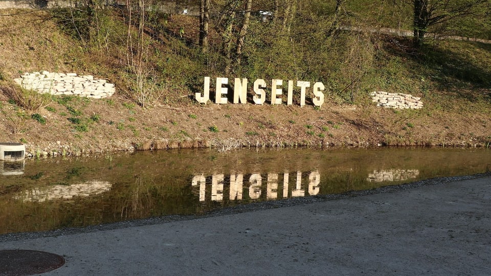 Holzbuchstaben bilden an einem Grashang das Wort Jenseits, davor ein Teich.