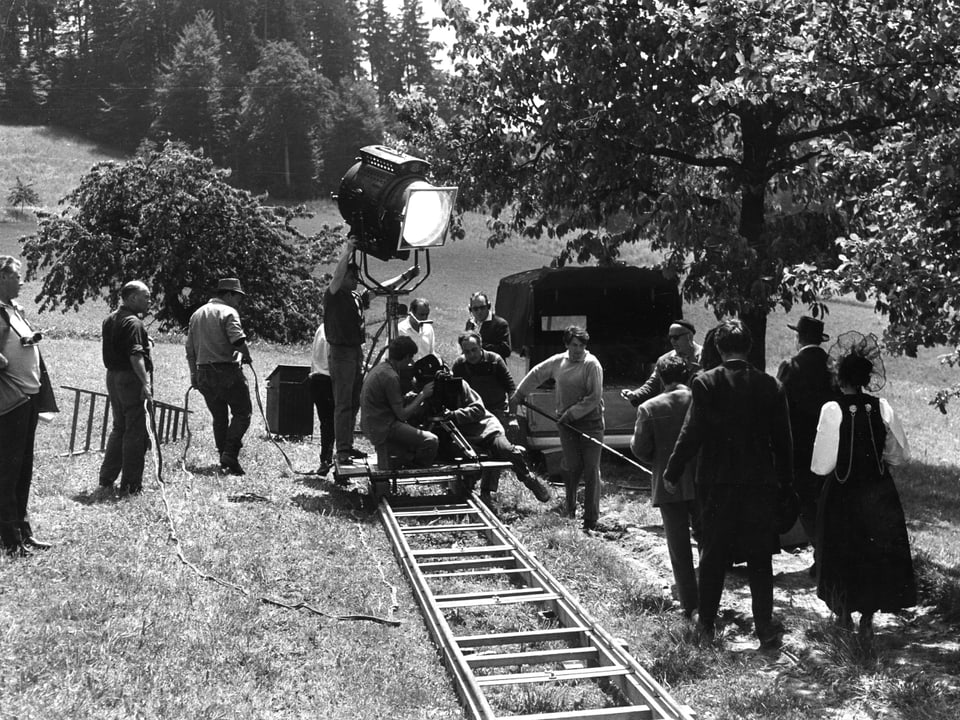 Kamerafahrt auf Schienen entlang eines Feldweges. Eine Schauspielerin und ein Schauspieler gehen den Weg entlang. Auf dem Kamerawagen steht ausserdem ein grosser Filmscheinwerfer.