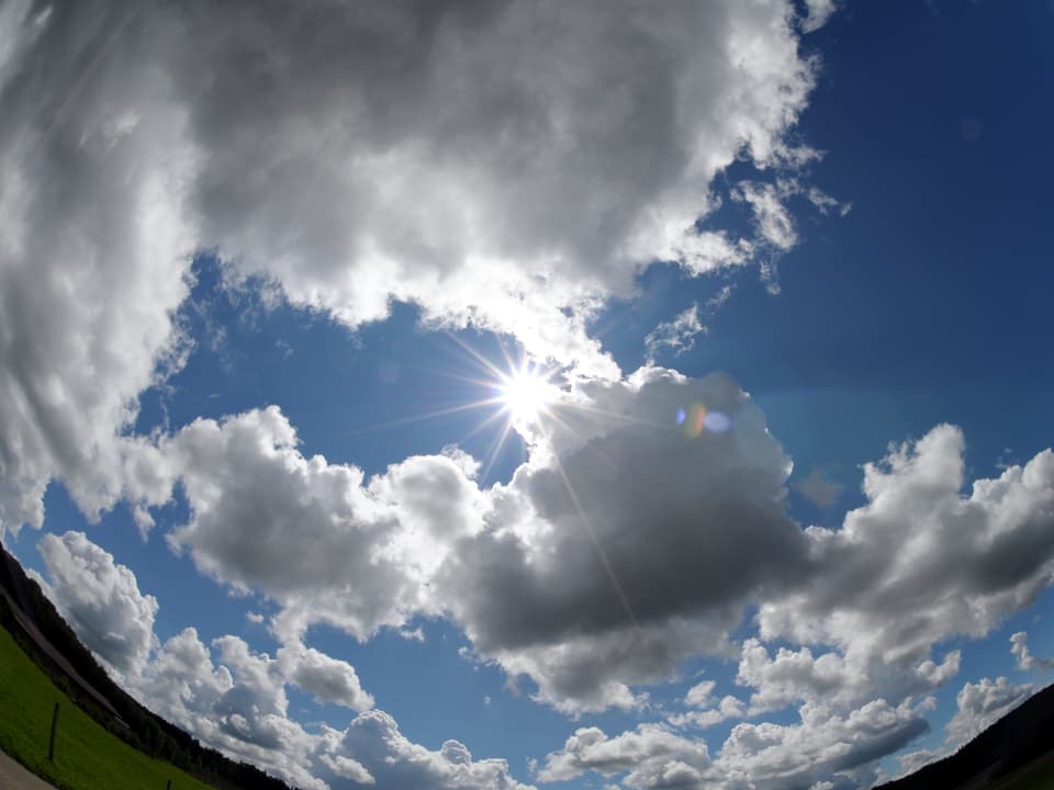 In der Mitte des Bildes die Sonne, rund herum blauer Himmel mit vielen Wolken. Das Foto ist mit einem Fischauge gemacht, am unteren Bildrand sieht man kreisförmige Konturen der Landschaft. 