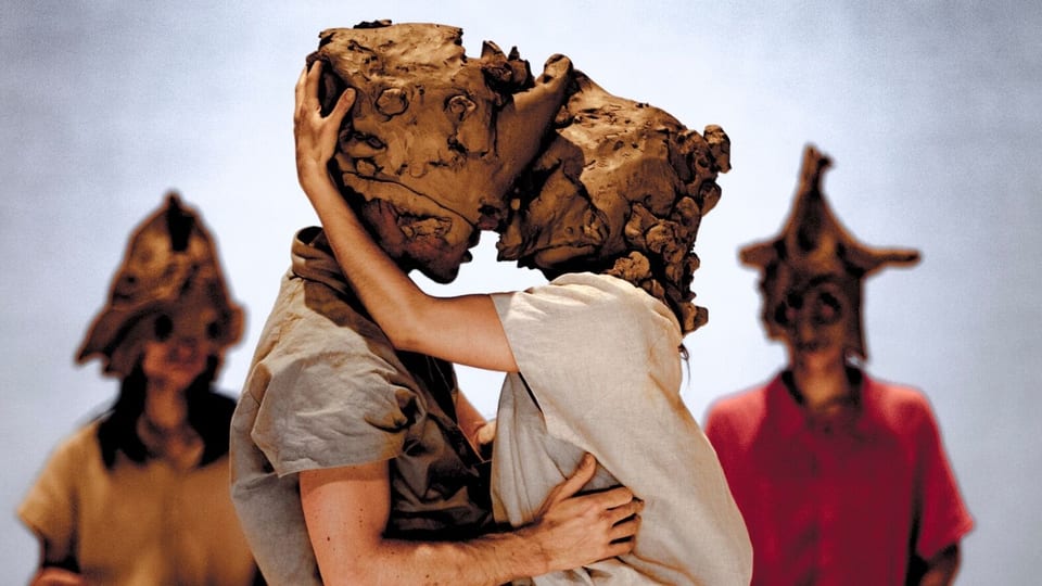 Zwei Tänzer mit Masken aus Ton küssen sich.