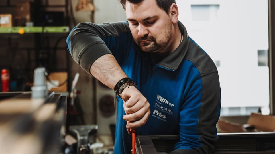 Mann mit dunklem Haar und kurzem Bart hält Werkzeug in der Hand, den Blick darauf gerichtet, steht in Werkstatt.