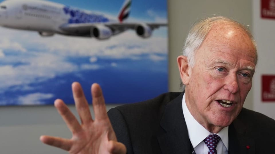 Tim Clark, der CEO von Emirates, gestikuliert in einem Stuhl sitzend vor einem Bild eines A380 in den Emirates-Farben.