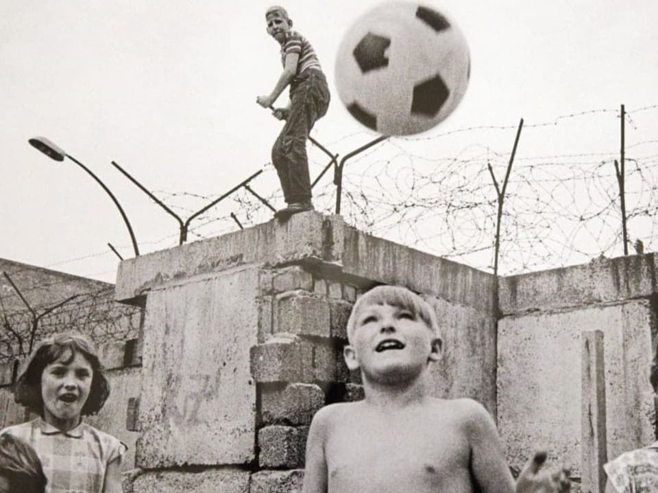 Black and white photo of three children, one of them raising his hands to catch a soccer ball in the air behind a wall.