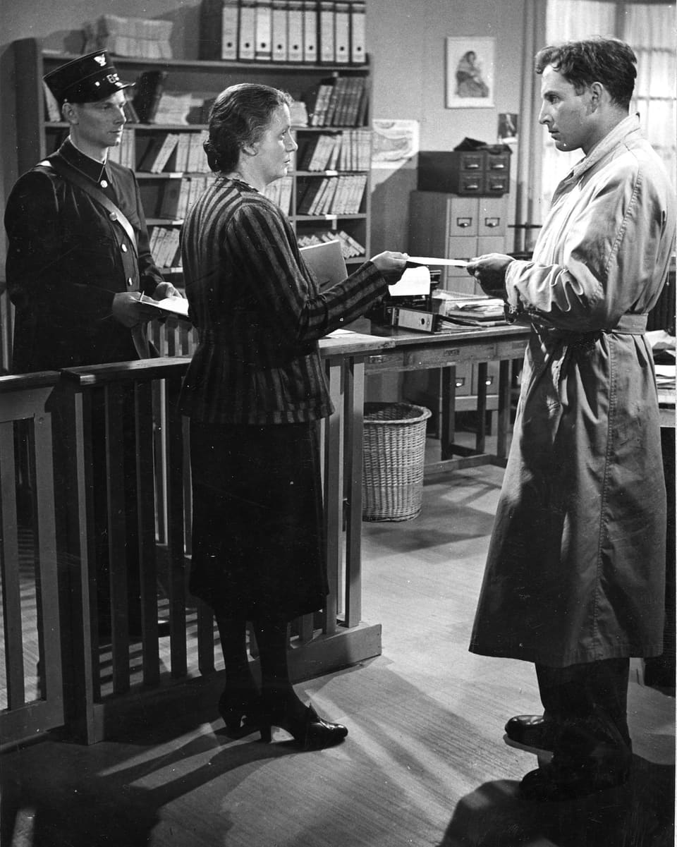 Zwei junge Männer und eine Frau stehen in einem Büro. Der Mann rechts überreicht der Frau in der Mitte einen Briefumschlag.