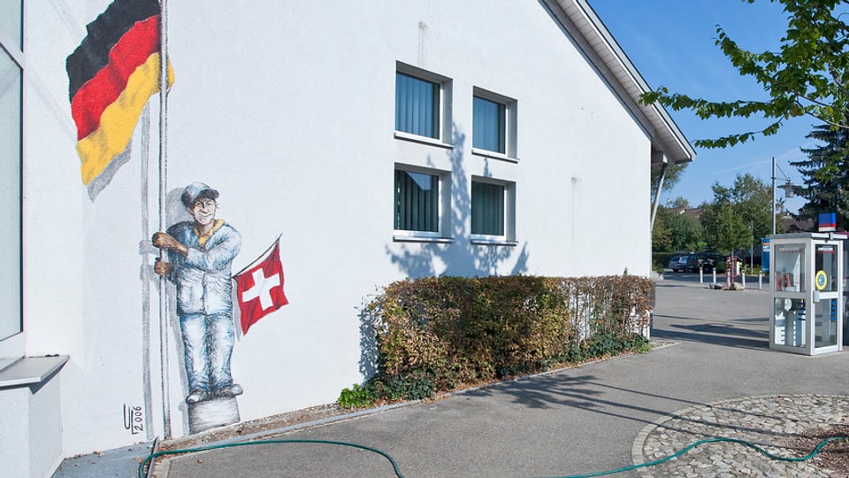 Eine Hausfassade, ein Mann aufgemalt, der eine grosse Deutschlandfahne und eine kleine Schweizfahne hält.