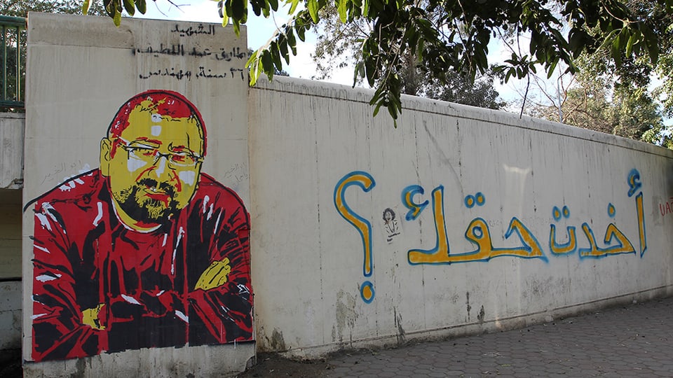 Abbildung eines Graffitos auf einer Mauer: Portrait eines Mannes mit Brille und Bart, der die Arme vor dem Körper verschrämkt hält. 