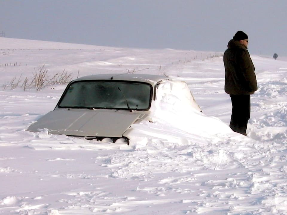 Die grossen Schneeverwehungen haben ein Auto komplett blockiert. Ein Mann steht vor dem Auto und starrt in die kalte Winterlandschaft.
