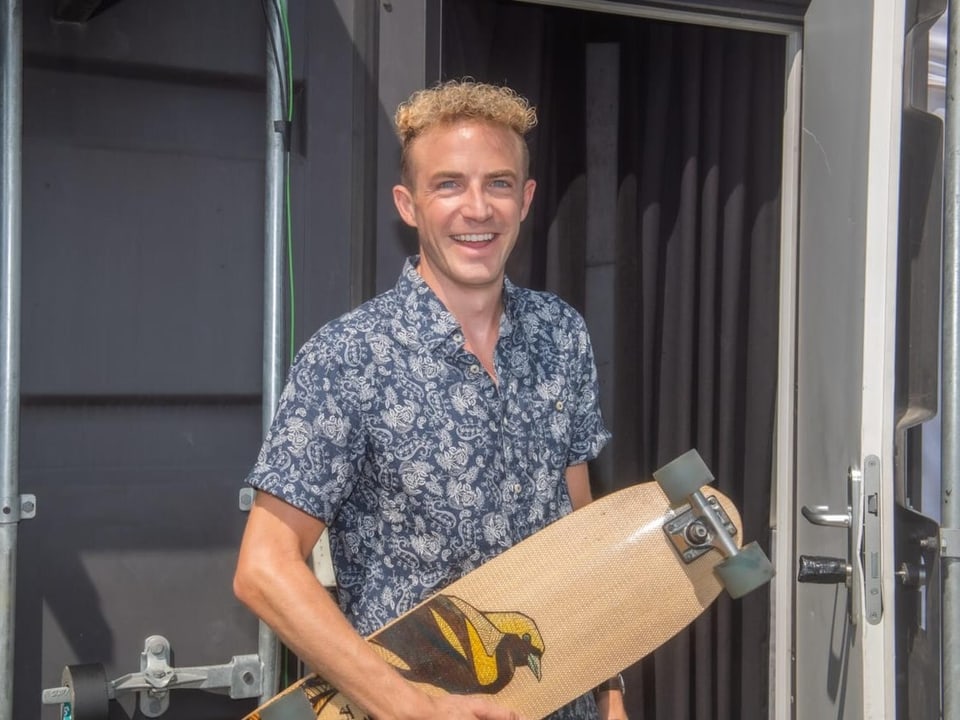 Ein Mann steht in einem farbigen Hemmli mit Rollbrett vor dem SRF 3-Sendestudio.