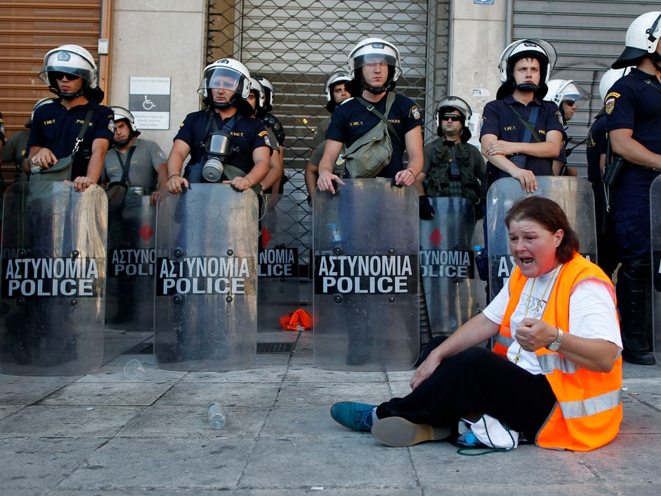 Eine Frau sitzt am Boden und ruft, hinter ihr stehen Polizisten in Kampfmontur.