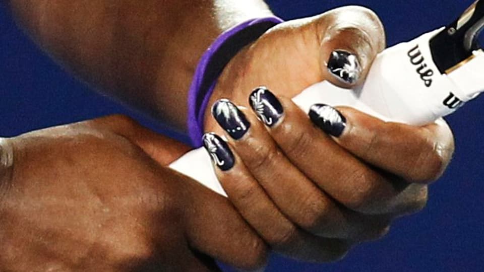 Serena Williams Hände halten ihr Racket. Ihre Fingernägel sind schwarz lackiert und haben ein silbriges Muster.