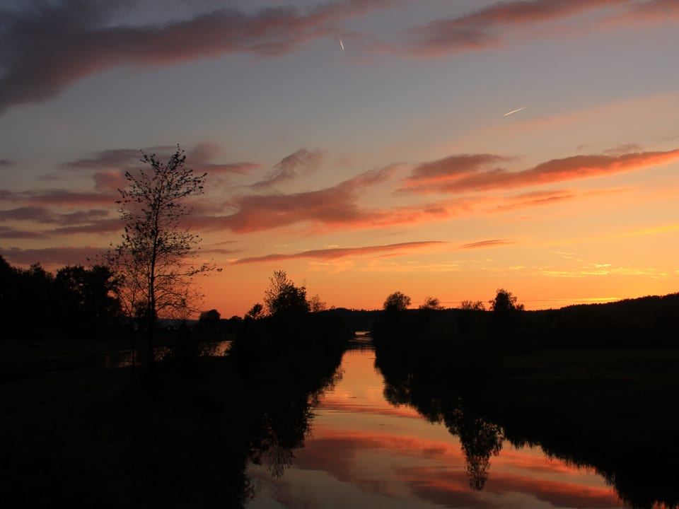 Das Bild ist in rosa und lila Farben gehalten. Man erkennt im Abendlicht nur Konturen von einem Fluss, Bäumen und Feldern. Am Himmel hat es ein paar wenige Wolken. 