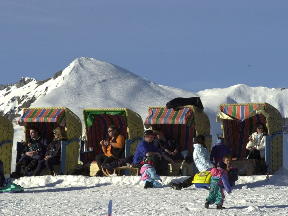 Strandkörbe stehen in Schnee. Im Pullover und mit Sonnenbrille ausgerüstet sitzen Wintersportbegeisterte in der Sonne. Der Himmel darüber ist strahlend blau.