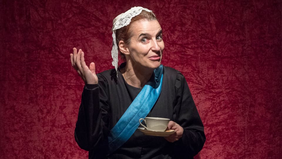 Eine Frau mit Kopfbedeckung gestikuliert mit einer Tasse in der Hand.
