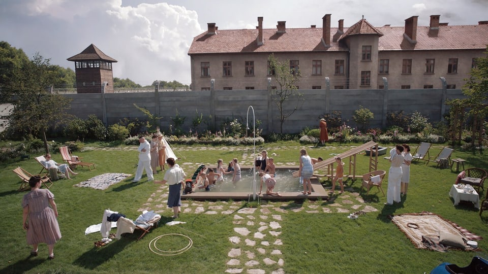 Liebevoll gestalteter Garten mit grünem Rasen, mittig baden Kinder in einem Pool. Dahinter eine Mauer mit Stacheldraht