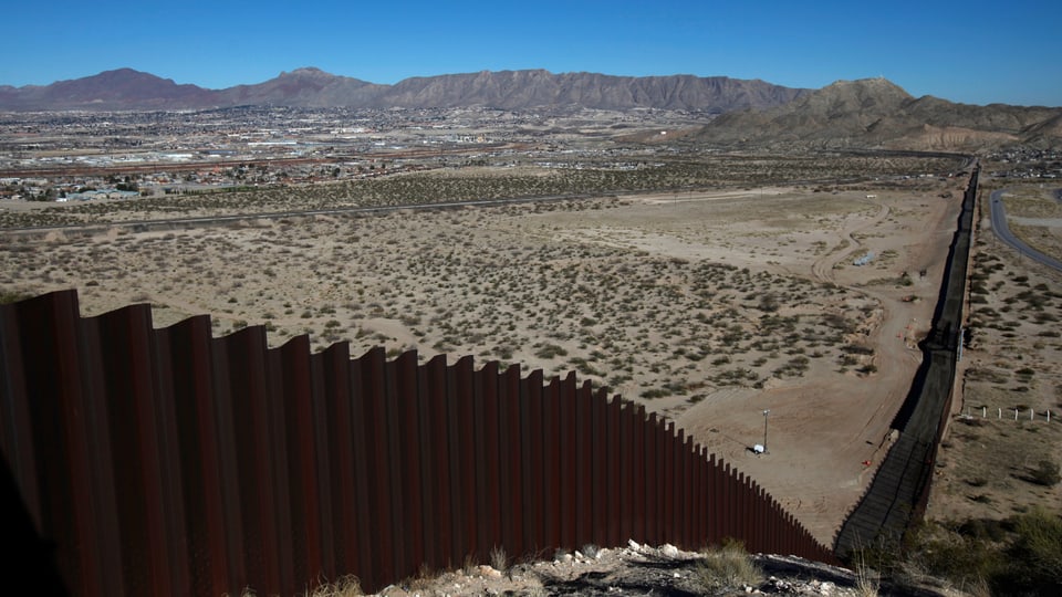 Grenzzaun zwischen den USA und Mexiko von einem Hügel aus in die Wüste herab fotografiert, im Hintergrund Berge