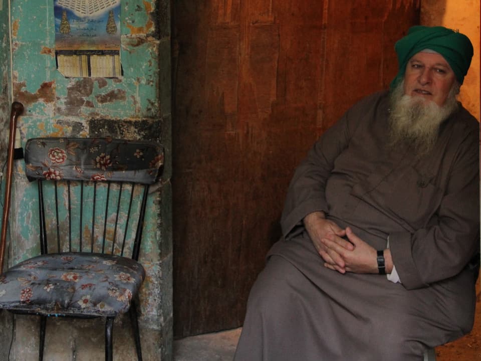 Ein älterer Moslem mit Bart sitzt vor einer Holztür.