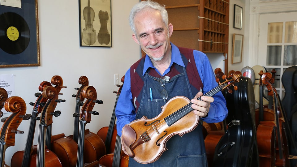 Roland Feller mit einer Geige in den Händen. Im Hintergrund Celli. 