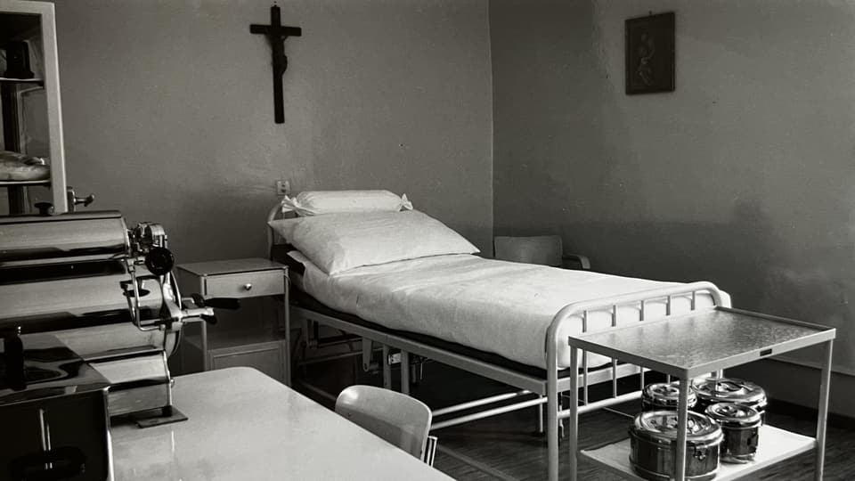 Schwarz-Weiss-Aufnahme eines Krankenzimmers mit Bett, Kreuz an der Wand und medizinischen Geräten.