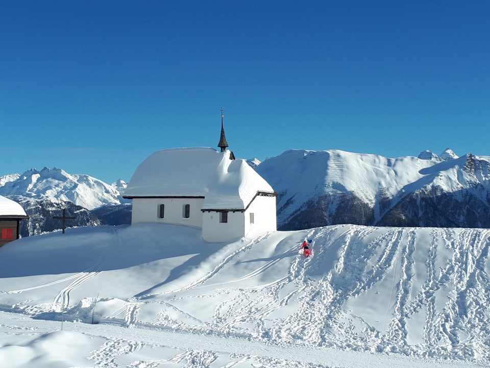 Winterlandschaft mit Kapelle auf einem Bergücke, überall liegt Schnee, der Himmel ist wolkenlos und  blau.