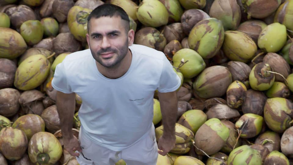 Gabriel Mascaro in weisser Kleidung auf einem Berg von Kokosnüssen. 