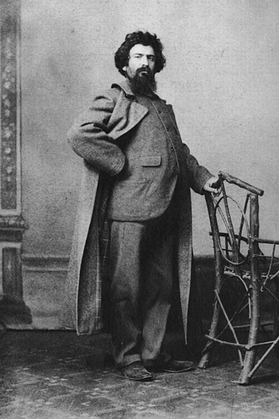 Giovanni Segantini, im Anzug, darüber ein Mantel, in herrschaftlicher Pose an einen Stuhl gestützt.