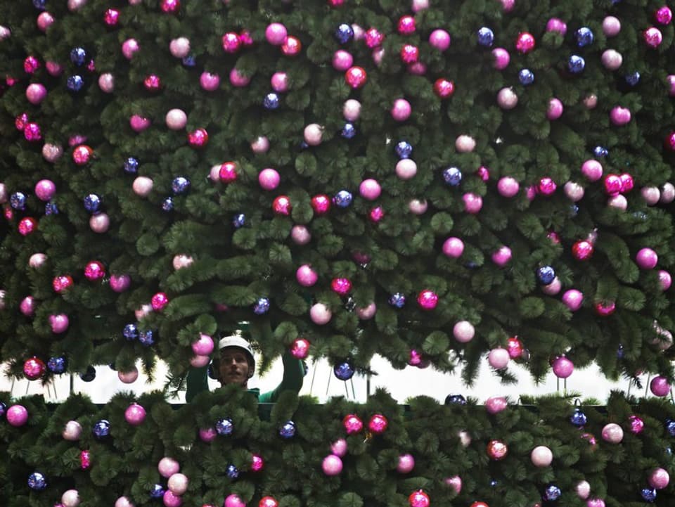 Blick durch einen grossen Weihnachtsbaum in Berlin mit vielen bunten Kugeln auf einen Mann, der die Kugeln aufhängt