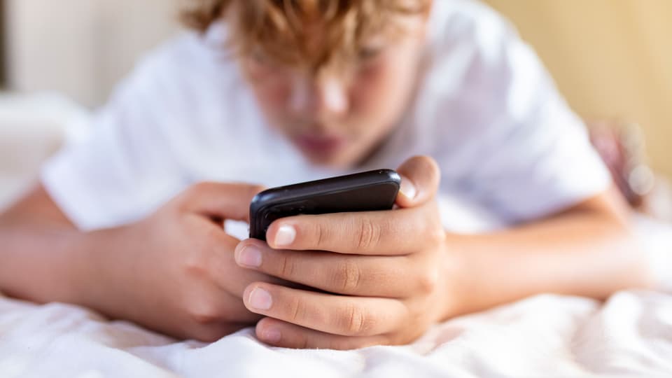 Ein Jugendlicher liegt auf dem Bett und hält ein Smartphone in seinen Händen.