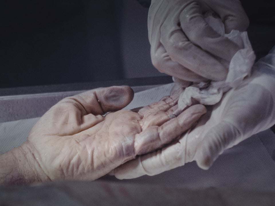Eine Hand mit Gummihandschuh reinigt die Hand eines Verstorbenen