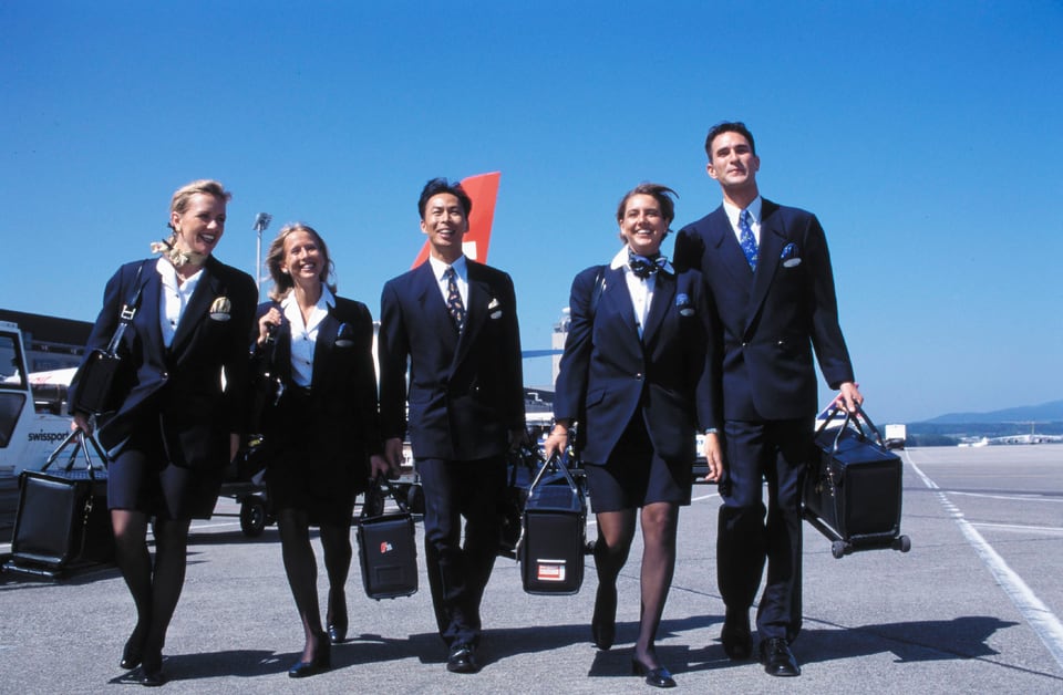 Fünf Frauen und Männer in dunkelblauen Uniformen, die Frauen mit Jupe, alle vor einem Flugzeug