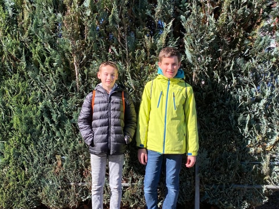 Zwei Jungs stehen vor einem grünen Strauch. Der eine trägt eine blau-graue, der andere eine hellgrüne Jacke.