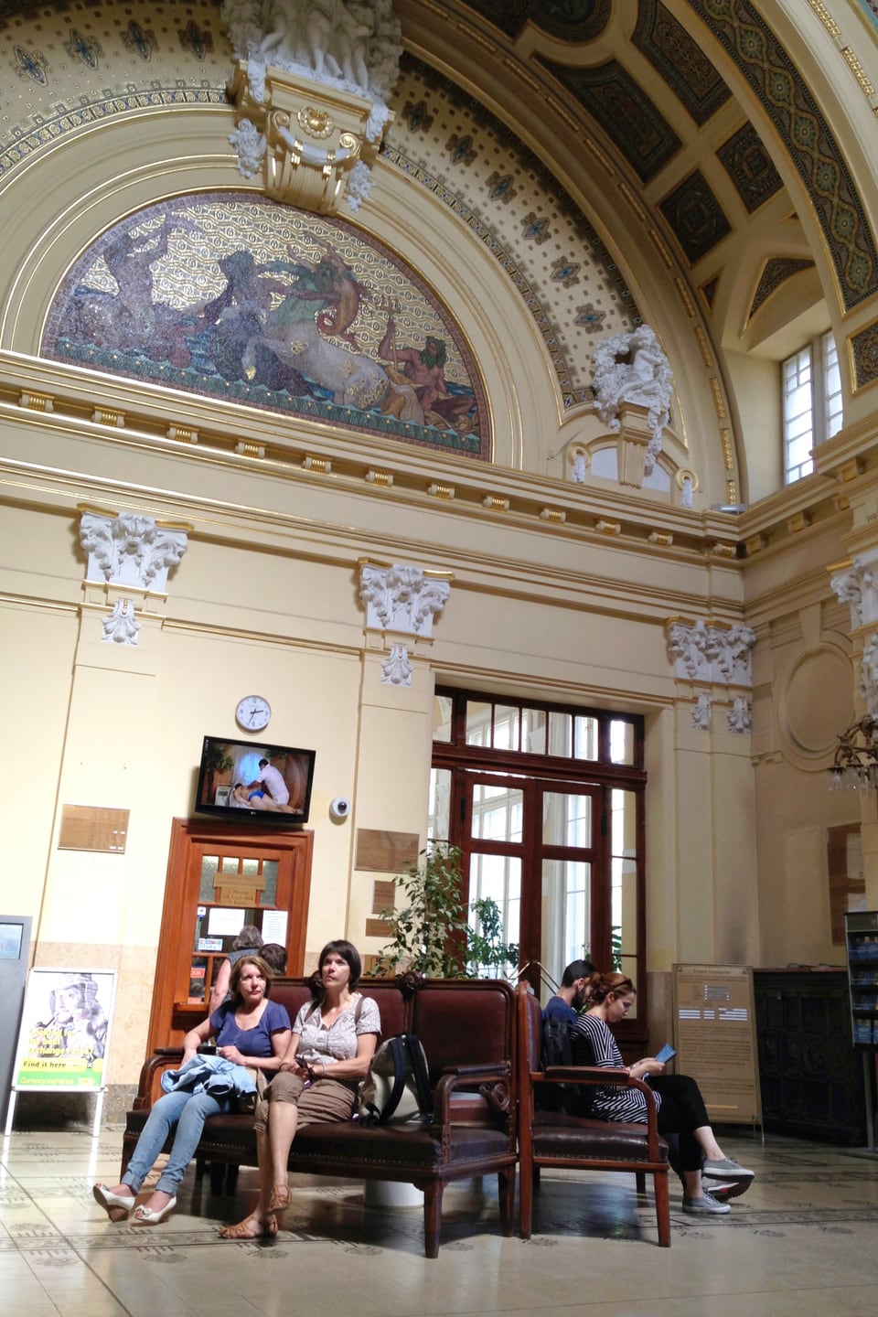 In einer geschmückten, hohen Eingangshalle sitzen ein paar Menschen auf Bänken.