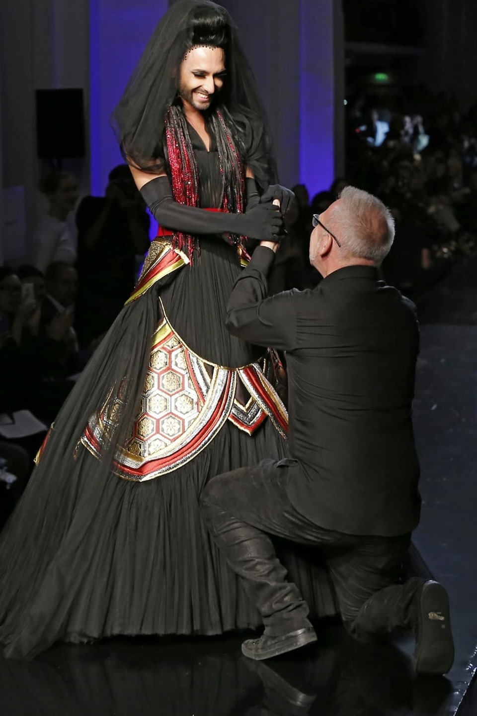 Jean-Pault Gaultier kniet vor Conchita Wurst, die ein schwarzes Brautkleid trägt.