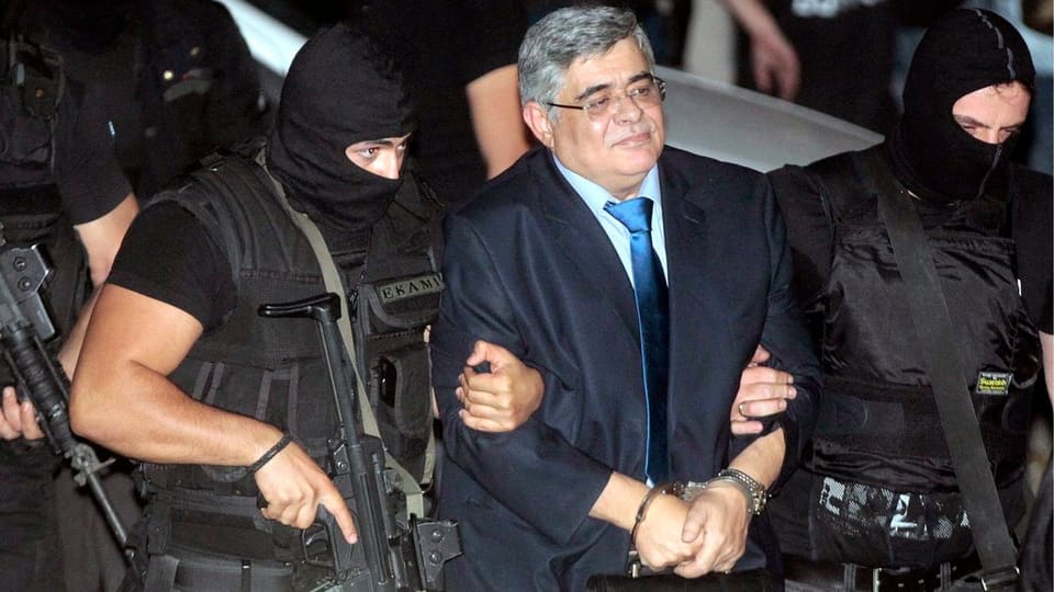 Nikos Michaloliakos wird mit Handschellen von bewaffneten Beamten in Roger-Staub-Mützen abgeführt.