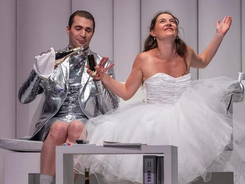 Mann in silbernem Anzug und Frau in weissem Brautkleid sitzen bei einer Theatervorstellung.