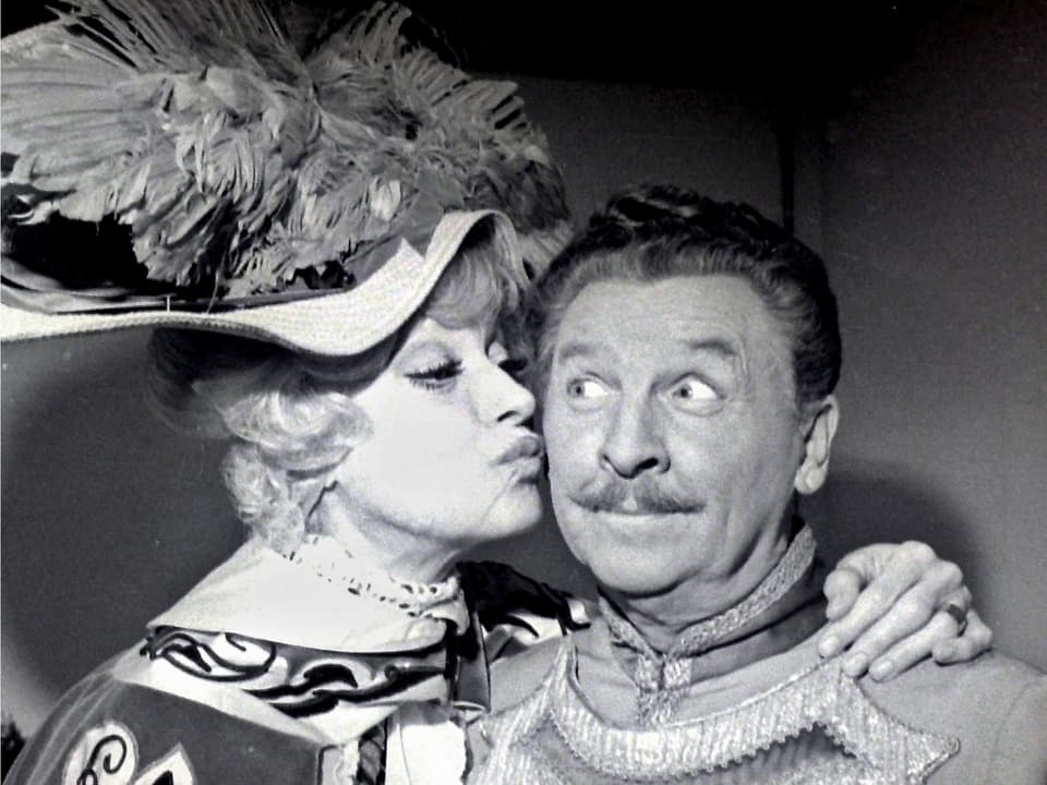 Carol küsst Eddie auf die Wange. Das Foto wurde Backstage während einer Vorstellung am Broadway aufgenommen. Beide Darsteller tragen ihre Kostüme: Carol Channing einen pompösen Federhut und Eddie Bracken eine Livrée.