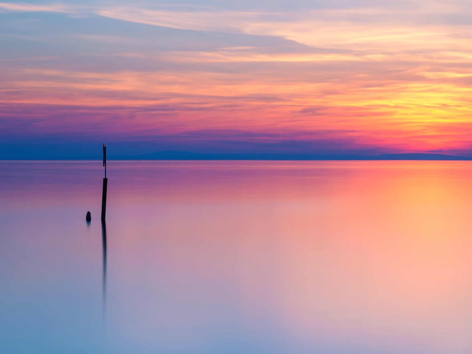 Pastellfarbiges Bild einer glatten Seeoberfläche mit einem Pfahl darin. Der Himmel ist milchig bewölkt und leuchtet in gelb bis rosa. Die Farben spiegeln sich im Wasser. 