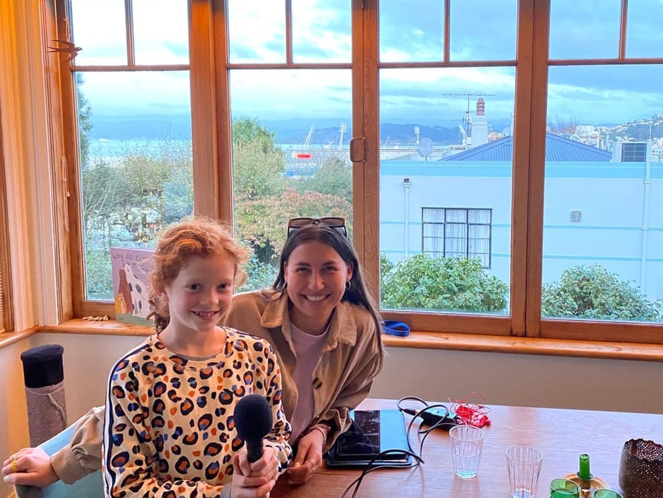 Clara und Anik sitzen am Esstisch im Wohnzimmer mit Aussicht auf Bäume, Häuser und den Hafen in Wellington.